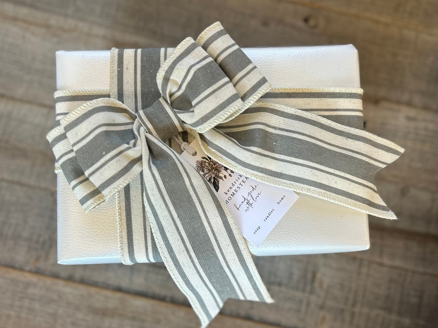 Farmhouse gift wrapping. white rectangular gift box wrapped with a wide cream and gray striped ribbon tied in a bow, with a small paper tag attached, placed on a rustic wood surface. Kendrick Homestead, 